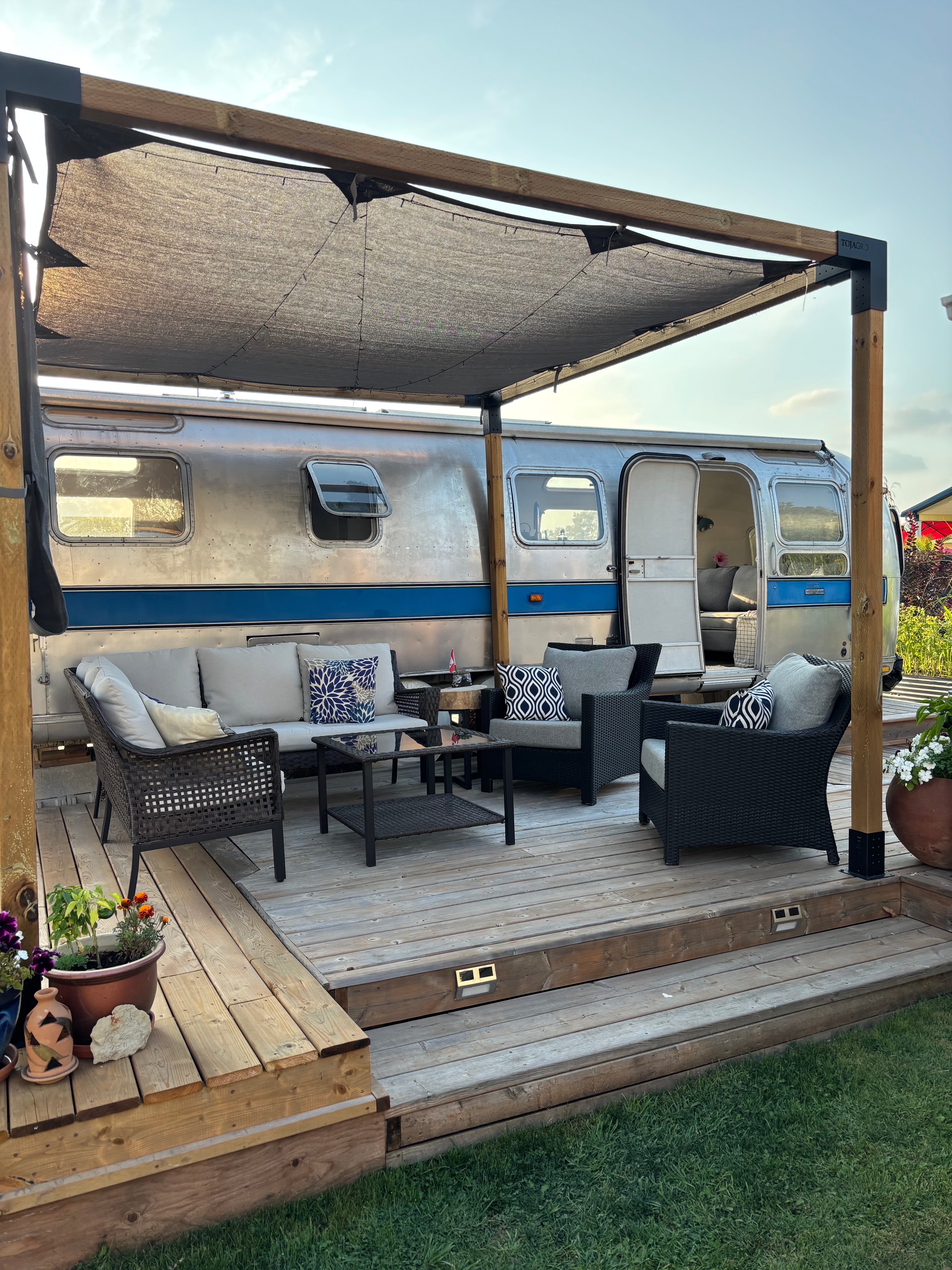 exterior side view of aluminum Airstream 1976 Excella Trailer parked beside wooden patio area with furnishings 