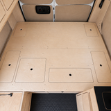 Overhead view of the integrated plywood bed platform in an Overland Interiors Low Roof Van Kit, showing multiple flush access panels, finger holes, rear-door insulation, upper cabinetry, and vinyl flooring
