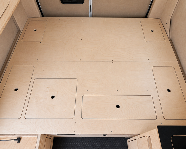 Overhead view of the integrated plywood bed platform in an Overland Interiors Low Roof Van Kit, showing multiple flush access panels, finger holes, rear-door insulation, upper cabinetry, and vinyl flooring