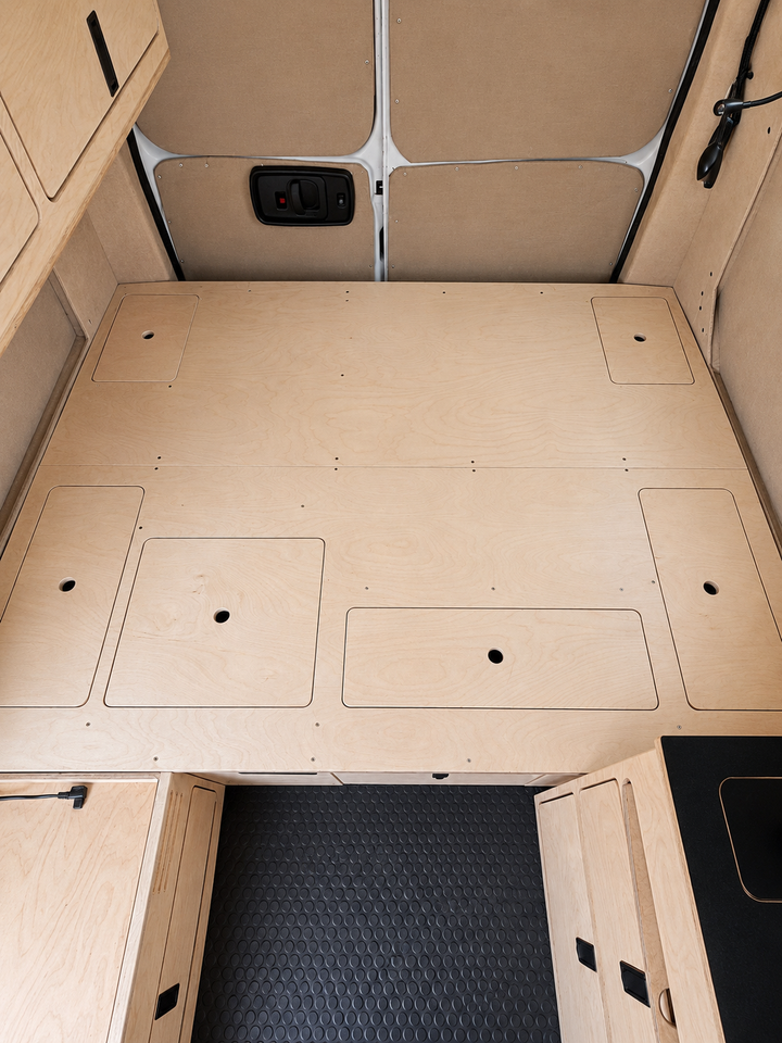 Overhead view of the integrated plywood bed platform in an Overland Interiors Low Roof Van Kit, showing multiple flush access panels, finger holes, rear-door insulation, upper cabinetry, and vinyl flooring