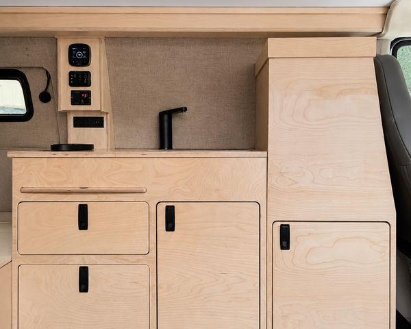 Front view of a 36-inch plywood camper van kitchenette with sink, faucet, electrical control panel, storage drawers, cabinet doors, and a tall utility cabinet in an Overland Interiors Low Roof Van Kit.