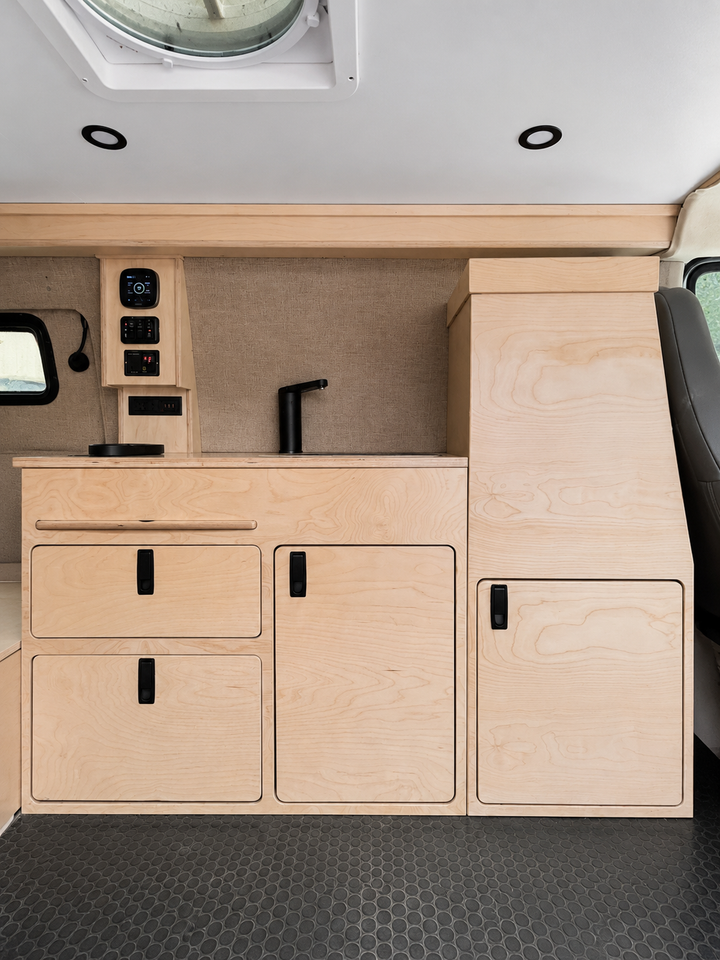 Front view of a 36-inch plywood camper van kitchenette with sink, faucet, electrical control panel, storage drawers, cabinet doors, and a tall utility cabinet in an Overland Interiors Low Roof Van Kit.