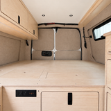 Rear view of the integrated plywood bed platform in an Overland Interiors Low Roof Van Kit, featuring hinged access panels, rear storage door, electrical outlets, upper cabinet, and interior lighting.