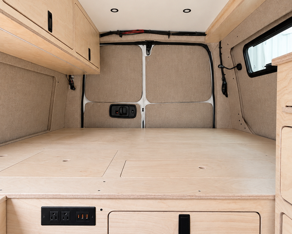 Rear view of the integrated plywood bed platform in an Overland Interiors Low Roof Van Kit, featuring hinged access panels, rear storage door, electrical outlets, upper cabinet, and interior lighting.