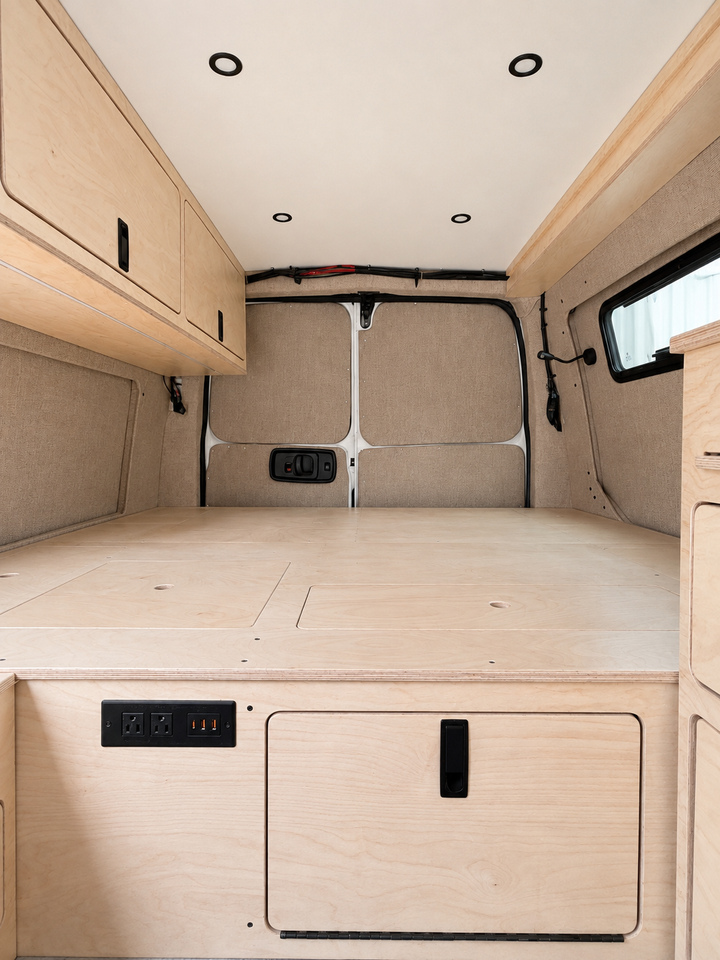 Rear view of the integrated plywood bed platform in an Overland Interiors Low Roof Van Kit, featuring hinged access panels, rear storage door, electrical outlets, upper cabinet, and interior lighting.