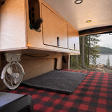 54-inch plywood upper cabinet with side panel installed above the bed in an Overland Interiors Low Roof Van Kit, shown with a Vancouver Island coastal forest view.