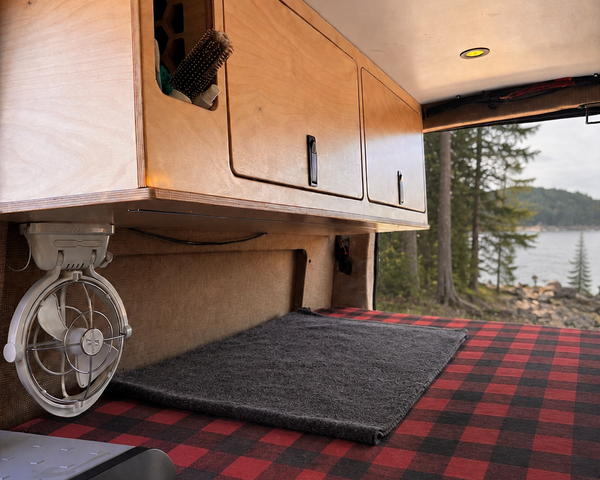 54-inch plywood upper cabinet with side panel installed above the bed in an Overland Interiors Low Roof Van Kit, shown with a Vancouver Island coastal forest view.