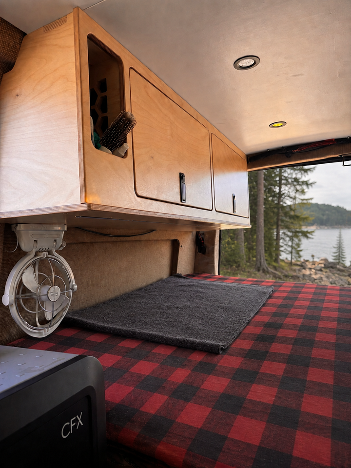 54-inch plywood upper cabinet with side panel installed above the bed in an Overland Interiors Low Roof Van Kit, shown with a Vancouver Island coastal forest view.